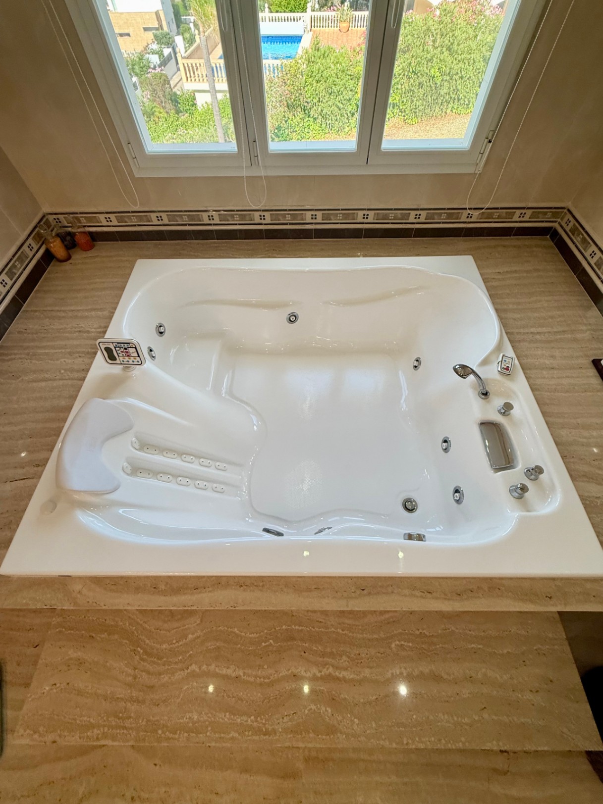 Baignoire d'hydromassage carrée encastrée dans un socle en travertin, équipée de commandes numériques et d'une fenêtre offrant une vue dégagée.