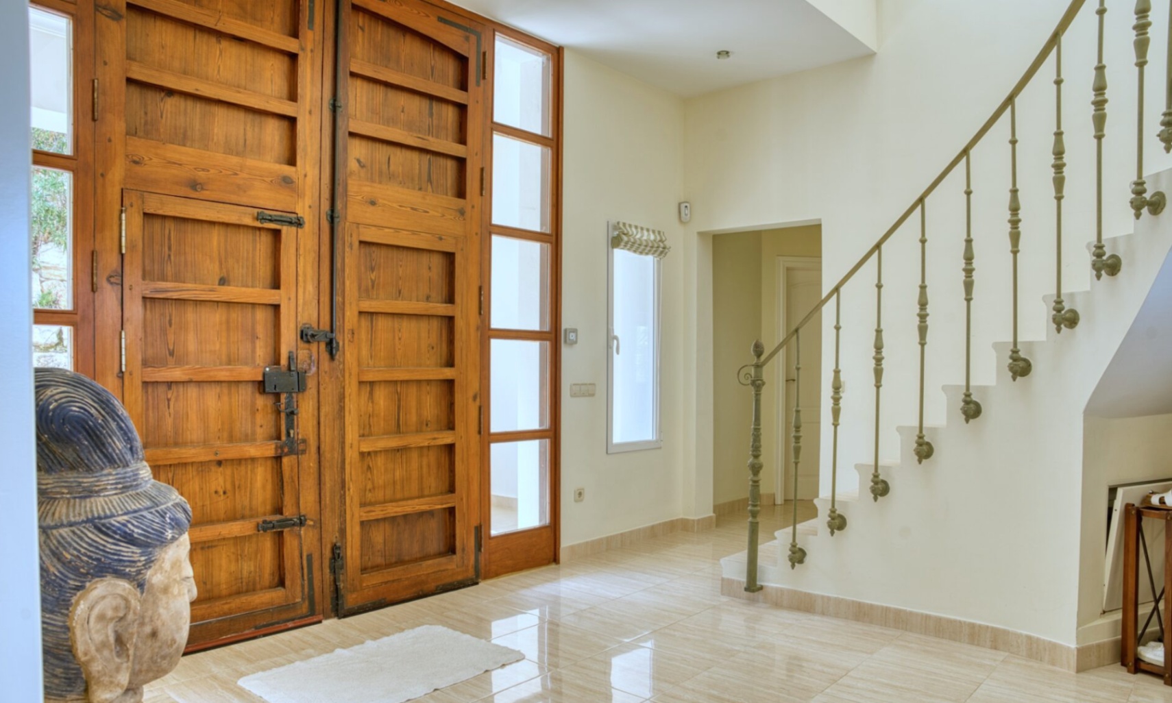 Hall d'entrée avec doubles portes en bois rustique, ferrures en fer forgé et sol en grès poli. Escalier avec rampe ornementale et lumière naturelle traversante.