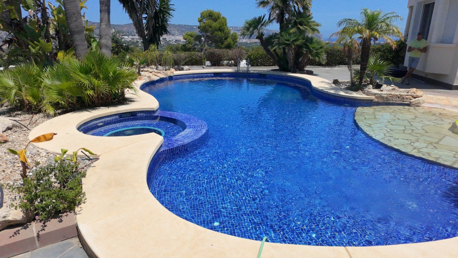 Piscine de forme libre avec mosaïque bleue et jacuzzi circulaire intégré, entourée d'une terrasse en pierre et de palmiers sous le ciel de Jávea.
