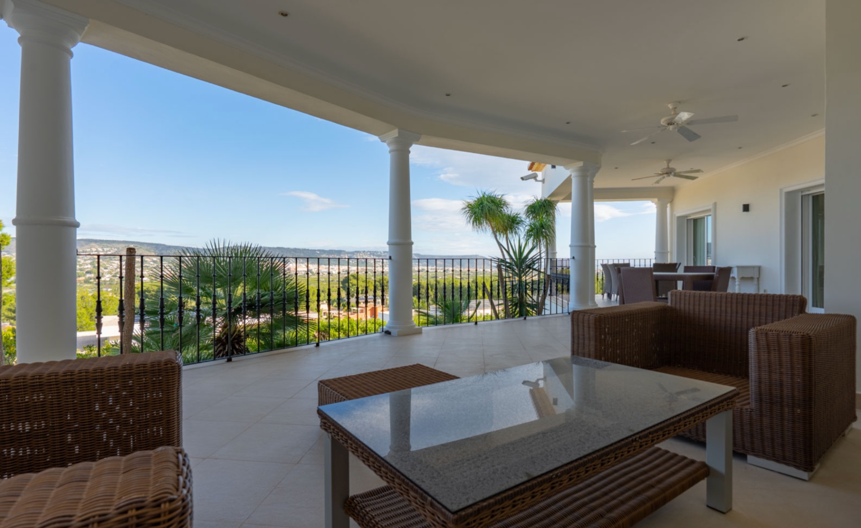 Terrasse couverte avec colonnes blanches et mobilier en rotin, offrant une vue panoramique sur la vallée. Équipée de ventilateurs de plafond et d'un éclairage intégré.