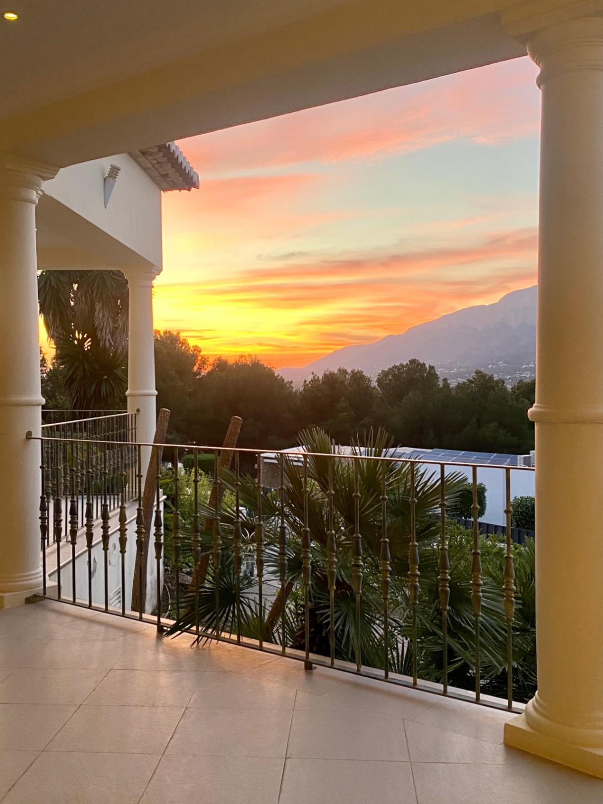Terrasse couverte avec colonnes classiques et garde-corps en fer, offrant une vue panoramique sur le coucher de soleil sur le Montgó et la végétation environnante.