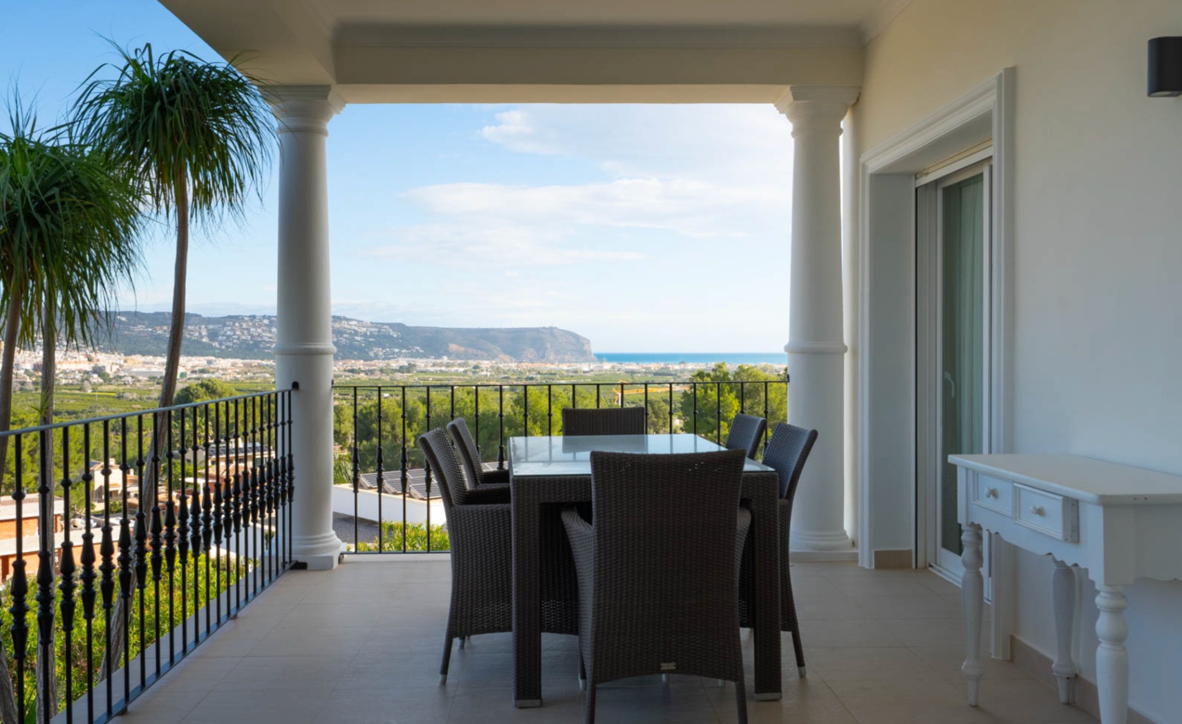 Terrasse couverte avec colonnes classiques, garde-corps en fer forgé et coin repas offrant une vue panoramique sur la mer Méditerranée et la vallée.