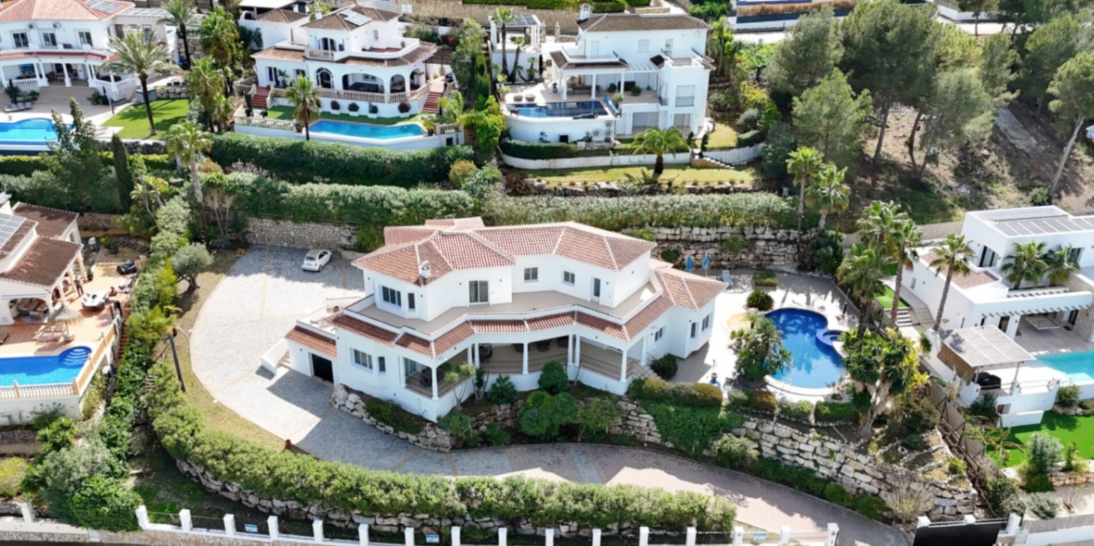 Vue aérienne de la villa principale avec toits en tuiles, large allée pavée, piscine privée entourée de palmiers et terrasses sur plusieurs niveaux intégrées au terrain.
