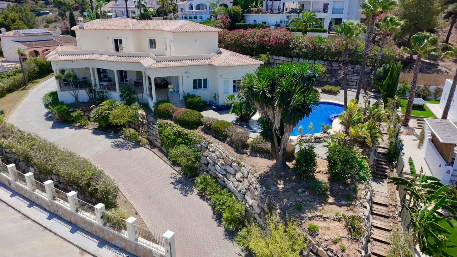 Vue aérienne de la villa révélant son architecture à deux niveaux, son allée pavée et sa piscine nichée au cœur d'un jardin paysager avec murs en pierre.