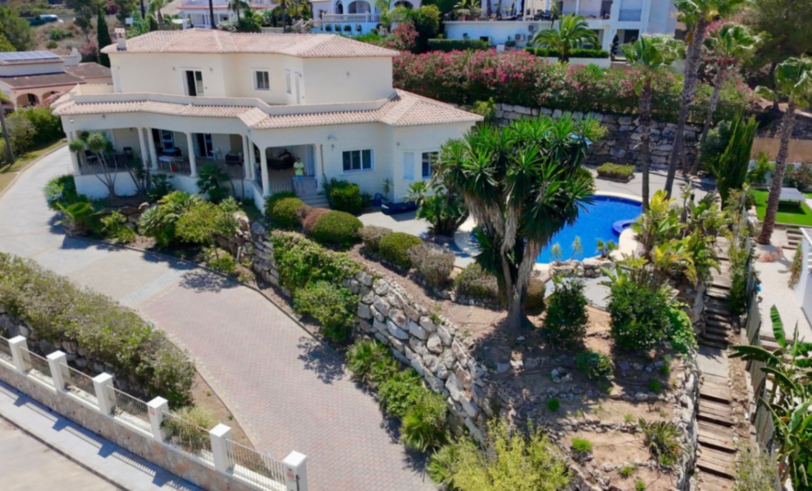 Vue extérieure de la villa avec façade blanche, toitures en tuiles, large allée pavée, murs de soutènement en pierre naturelle et jardins paysagers avec piscine.