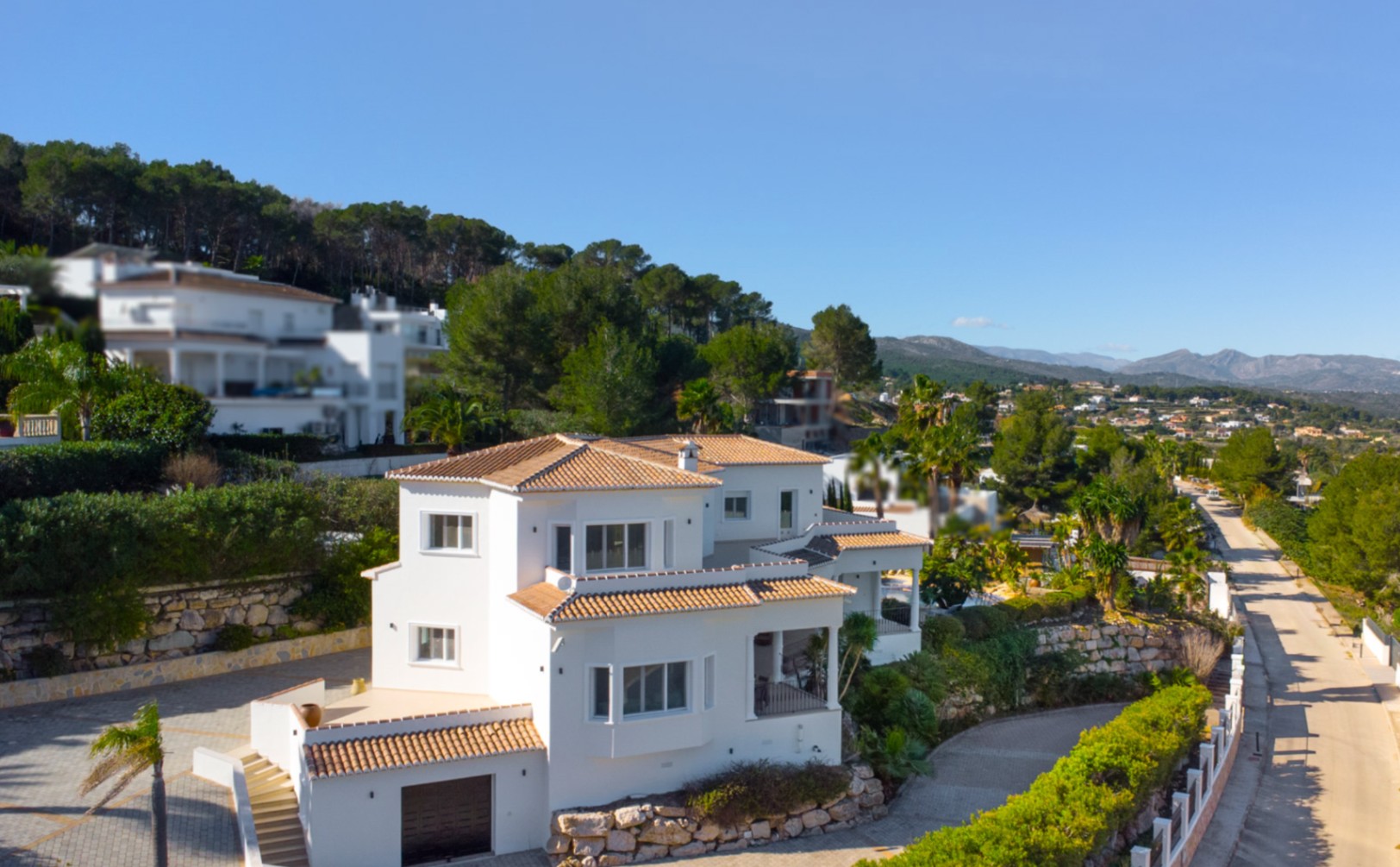 Vue extérieure de la villa méditerranéenne aux façades blanches et toits de tuiles, nichée sur un terrain en pente avec des murs en pierre naturelle et un panorama montagneux.